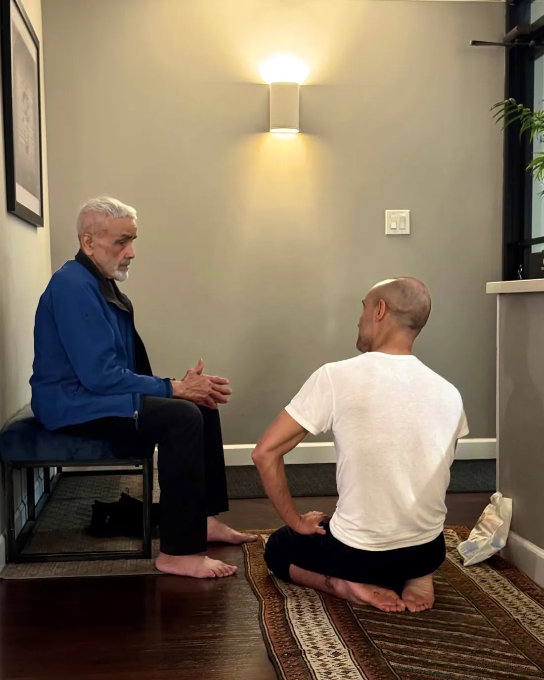Andrei Ram, sentado en el suelo sobre una alfombra, conversa con Sri Dharma Mittra, sentado frente a él en un banco, en la escuela de Dharma en Nueva York